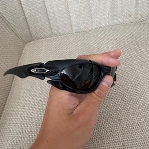 May include: Black sunglasses with a modern design. The sunglasses have a curved frame and dark lenses. The Oakley logo is visible on the temple. The sunglasses are held in a hand.