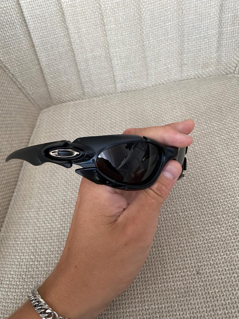 May include: Black sunglasses with a modern design. The sunglasses have a curved frame and dark lenses. The Oakley logo is visible on the temple. The sunglasses are held in a hand.
