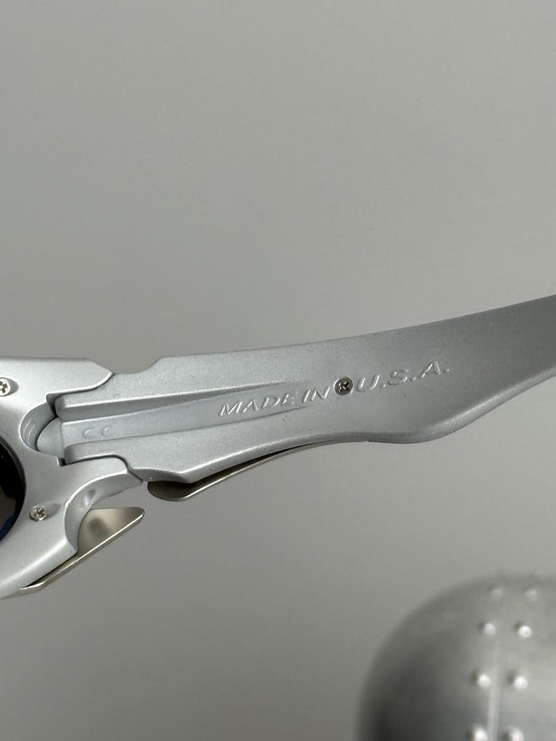 May include: Close-up of a silver-colored sunglasses arm. The text "MADE IN U.S.A." is engraved on the arm. The sunglasses arm is made of a metallic material and has a sleek, modern design. The background is a solid gray.