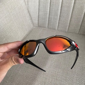 Op de afbeelding: Zwarte zonnebril met oranje en rode glazen. De zonnebril heeft een sportief design met een zwart montuur en een rode accentkleur aan de zijkant. De glazen hebben een verloop van oranje naar rood.