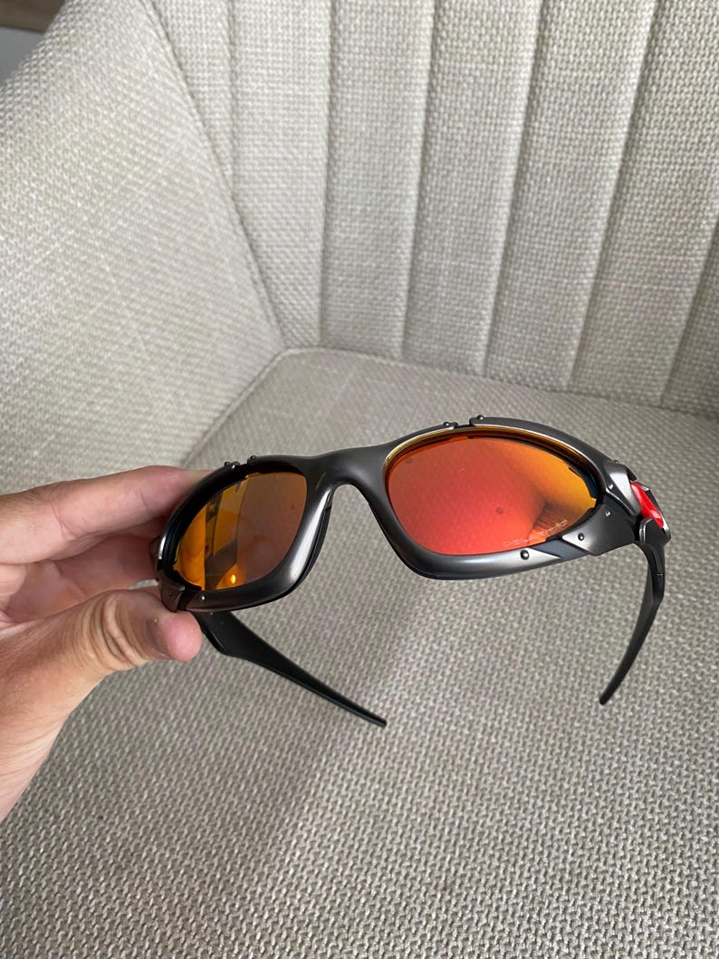 Op de afbeelding: Zwarte zonnebril met oranje en rode glazen. De zonnebril heeft een sportief design met een zwart montuur en een rode accentkleur aan de zijkant. De glazen hebben een verloop van oranje naar rood.