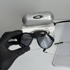 K&ouml;nnte beinhalten: Schwarze Sonnenbrille mit runden Gl&auml;sern und einem schwarzen Rahmen. Die Sonnenbrille wird in einer schwarz behandschuhten Hand gehalten. Ein silbernes Etui und eine Schachtel mit dem Text "SCIENCE AND ART" befinden sich im Hintergrund.