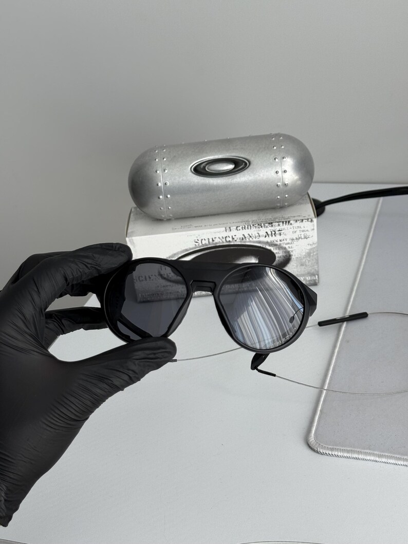 K&ouml;nnte beinhalten: Schwarze Sonnenbrille mit runden Gl&auml;sern und einem schwarzen Rahmen. Die Sonnenbrille wird in einer schwarz behandschuhten Hand gehalten. Ein silbernes Etui und eine Schachtel mit dem Text "SCIENCE AND ART" befinden sich im Hintergrund.