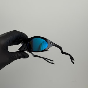 May include: Black sunglasses with blue mirrored lenses. The sunglasses have a unique, angular frame design. The lenses reflect a bright blue color. The arms of the sunglasses have a distinctive, curved shape.