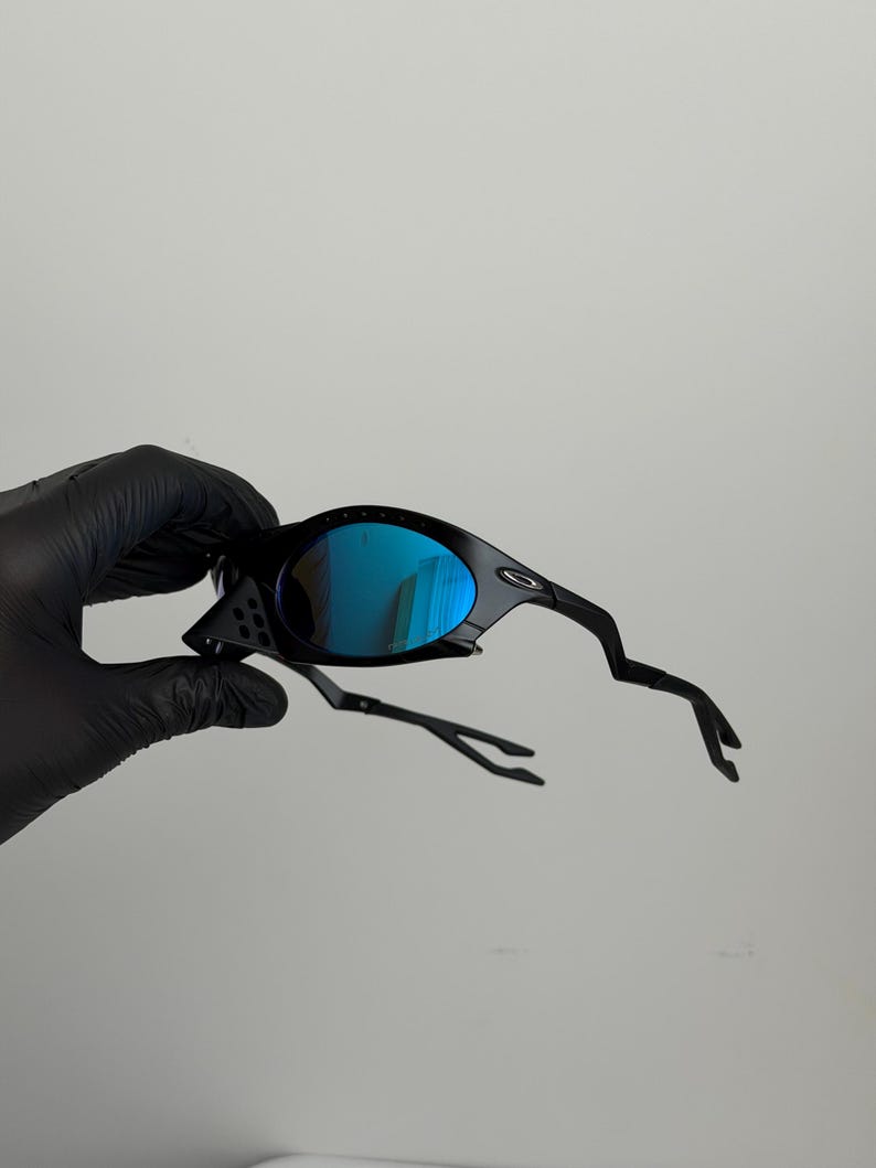 May include: Black sunglasses with blue mirrored lenses. The sunglasses have a unique, angular frame design. The lenses reflect a bright blue color. The arms of the sunglasses have a distinctive, curved shape.