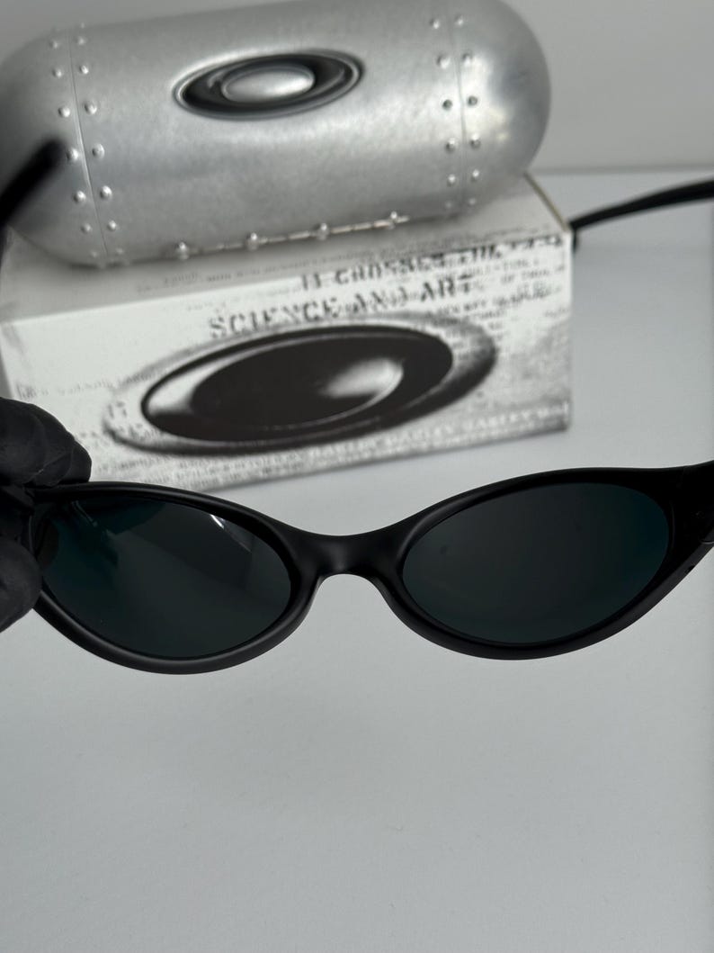 May include: Black oval-framed sunglasses with dark lenses. The image shows the sunglasses against a white backdrop, with a silver case and a box with the text "SCIENCE AND ART".
