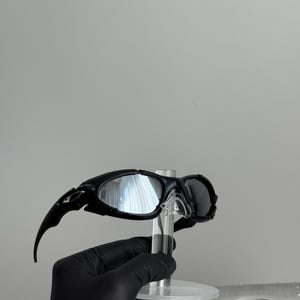 May include: Black sunglasses with a sleek, modern design. The sunglasses have a curved frame and dark lenses. The arms of the sunglasses extend outward with a pointed tip. The sunglasses are displayed on a clear stand.