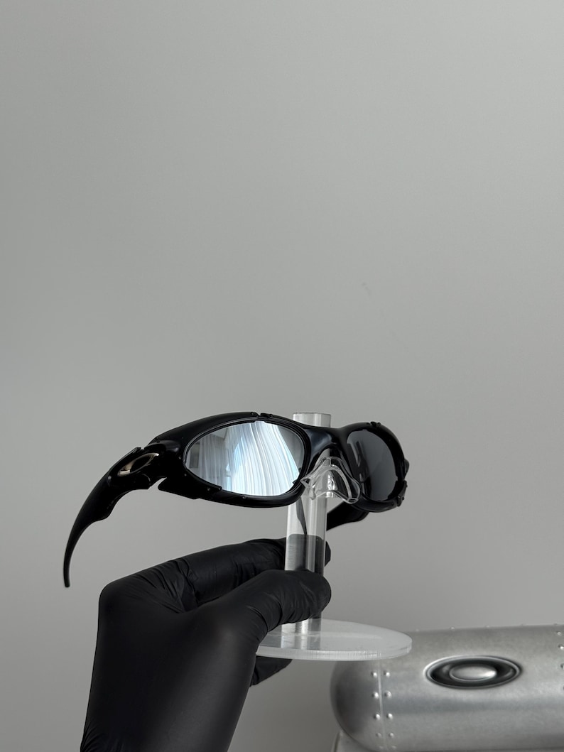 May include: Black sunglasses with a sleek, modern design. The sunglasses have a curved frame and dark lenses. The arms of the sunglasses extend outward with a pointed tip. The sunglasses are displayed on a clear stand.