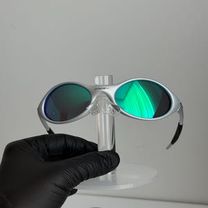 May include: Silver sunglasses with green lenses. The glasses are displayed on a clear stand, held by a gloved hand. The lenses reflect bright green light.