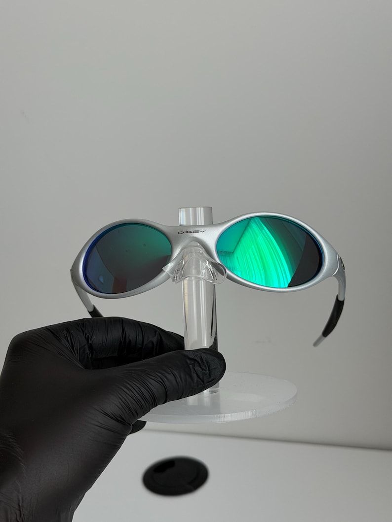 May include: Silver sunglasses with green lenses. The glasses are displayed on a clear stand, held by a gloved hand. The lenses reflect bright green light.