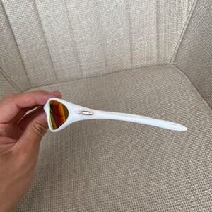 Puede incluir: Gafas de sol blancas con lentes de color naranja. Las gafas tienen una montura curvada y un peque&ntilde;o emblema plateado en la patilla. Las varillas son largas y afiladas, y la est&eacute;tica general es deportiva.