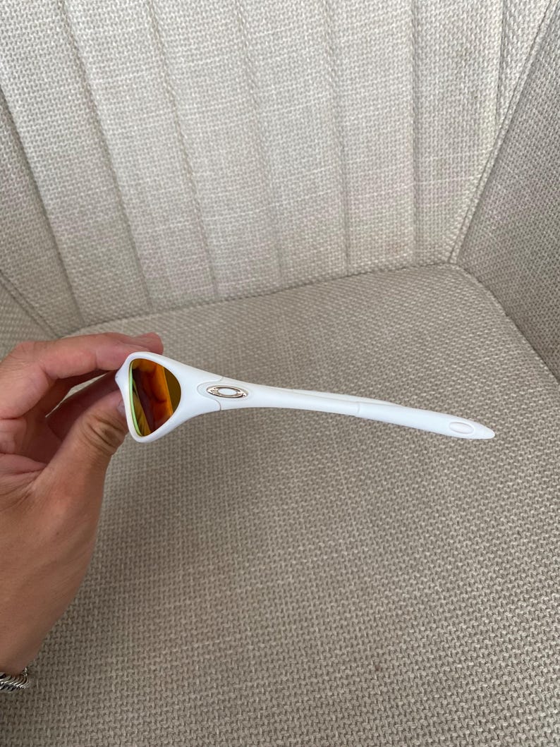 Puede incluir: Gafas de sol blancas con lentes de color naranja. Las gafas tienen una montura curvada y un peque&ntilde;o emblema plateado en la patilla. Las varillas son largas y afiladas, y la est&eacute;tica general es deportiva.