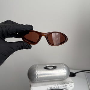 May include: Brown sunglasses with a matching case and box. The sunglasses have a curved frame and lenses. The case is silver and cylindrical. The box has the text "SCIENCE AND ART" and a logo.