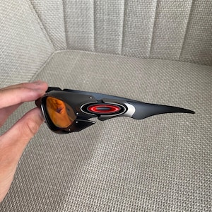 Op de afbeelding: Zwarte en grijze sportzonnebril met oranje glazen. De zonnebril heeft een strak ontwerp met een rode en zilveren accent op het montuur. De glazen zijn fel oranje en contrasteren met het donkere montuur. Het montuur is gemaakt van duurzaam materiaal.