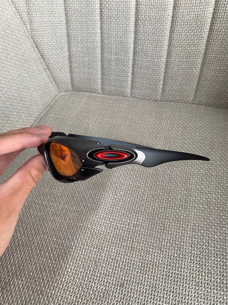Op de afbeelding: Zwarte en grijze sportzonnebril met oranje glazen. De zonnebril heeft een strak ontwerp met een rode en zilveren accent op het montuur. De glazen zijn fel oranje en contrasteren met het donkere montuur. Het montuur is gemaakt van duurzaam materiaal.