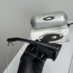 K&ouml;nnte beinhalten: Schwarze Sonnenbrille mit schlankem Design, gehalten von einer behandschuhten Hand. Die Brille hat eine moderne Form und wird mit einem silbernen Etui und einer Box mit dem Text "SCIENCE AND ART" kombiniert.