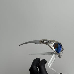 May include: Silver and blue sunglasses with a futuristic design. The sunglasses have a curved frame and blue lenses. The image shows the sunglasses on a clear stand, held by a gloved hand.