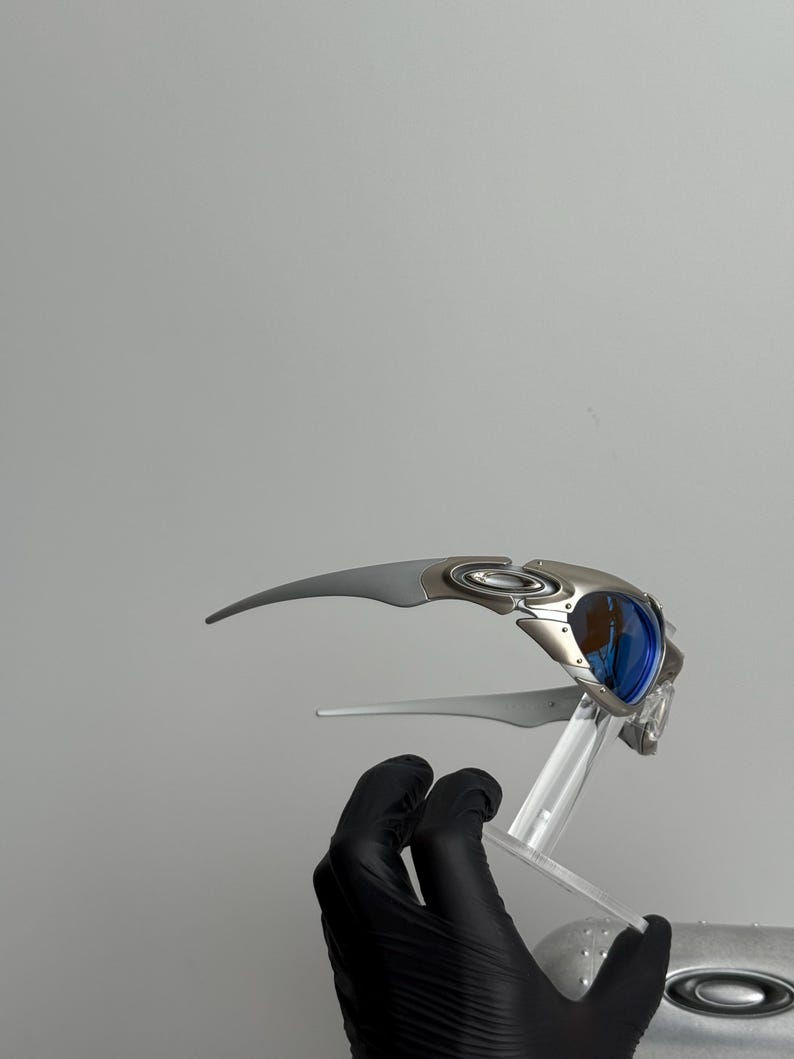 May include: Silver and blue sunglasses with a futuristic design. The sunglasses have a curved frame and blue lenses. The image shows the sunglasses on a clear stand, held by a gloved hand.