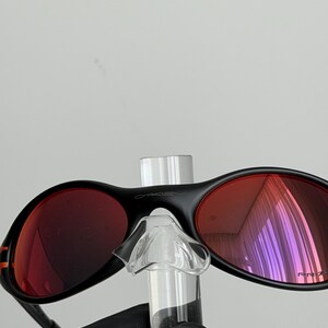 May include: Black Oakley sunglasses with red lenses. The sunglasses have a sleek, modern design with a black frame and a small orange stripe on the side. The lenses are a vibrant red color, and the Oakley logo is visible on the top of the frame.