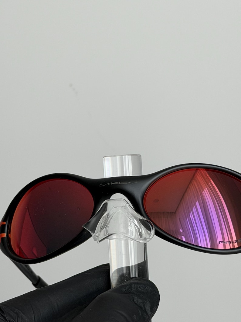 May include: Black Oakley sunglasses with red lenses. The sunglasses have a sleek, modern design with a black frame and a small orange stripe on the side. The lenses are a vibrant red color, and the Oakley logo is visible on the top of the frame.