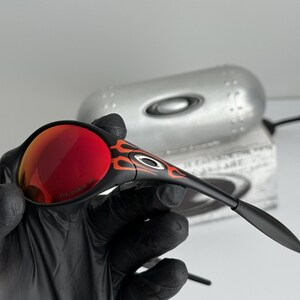 May include: Black sunglasses with red lenses and orange flame accents. The sunglasses have a sleek, modern design. The lenses are a vibrant red color, and the frames are black.