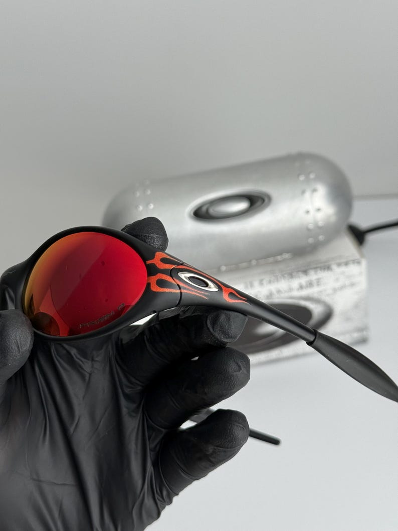 May include: Black sunglasses with red lenses and orange flame accents. The sunglasses have a sleek, modern design. The lenses are a vibrant red color, and the frames are black.