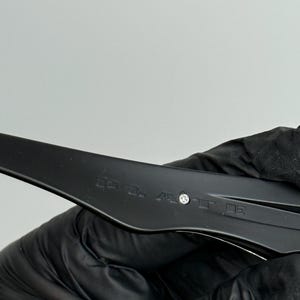 May include: Close-up of a black plastic object with text and a small silver rivet. The object is held by a gloved hand against a white background. The text is blurry.