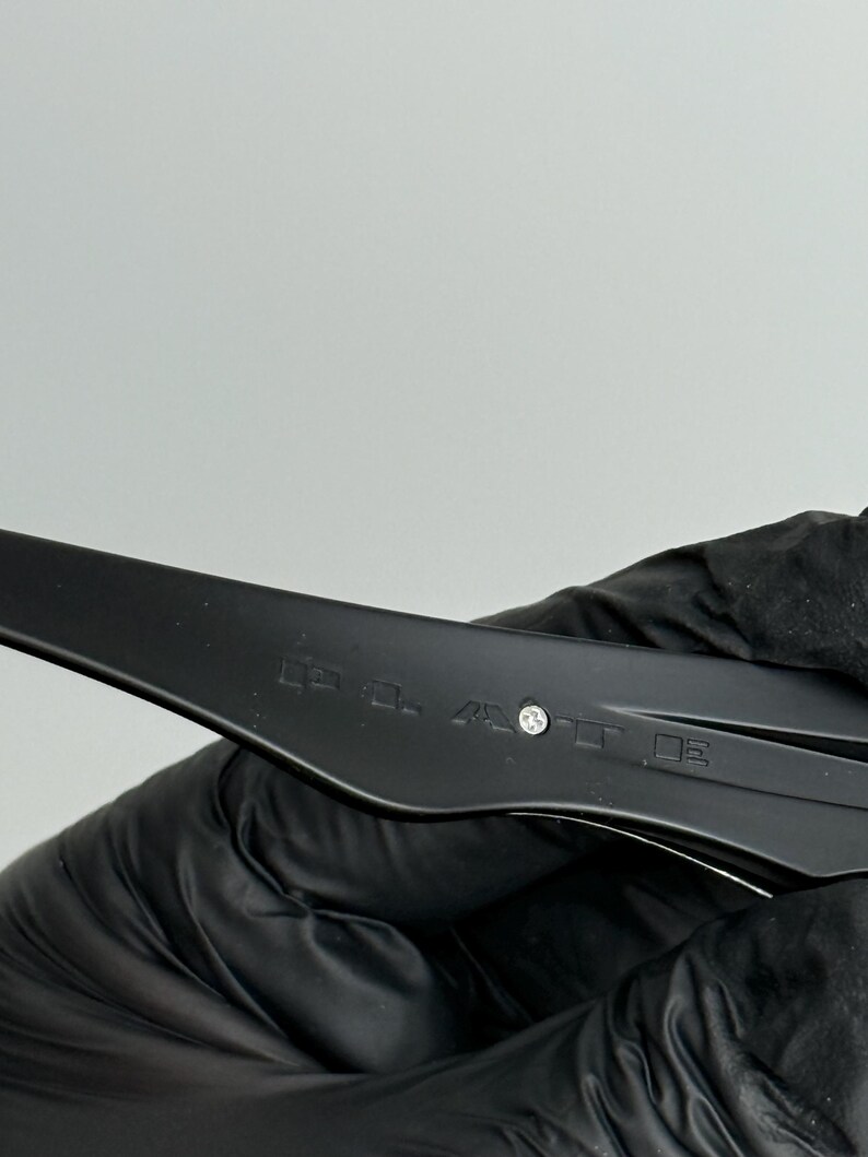 May include: Close-up of a black plastic object with text and a small silver rivet. The object is held by a gloved hand against a white background. The text is blurry.