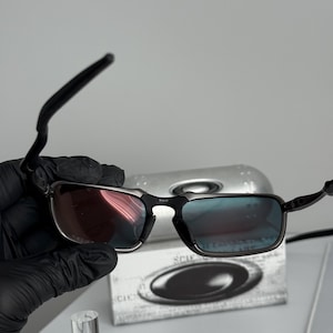 May include: Black rectangular sunglasses with a silver frame and gradient lenses. The lenses transition from red to blue. The sunglasses are held in a black gloved hand. A silver case with the brand name is in the background.