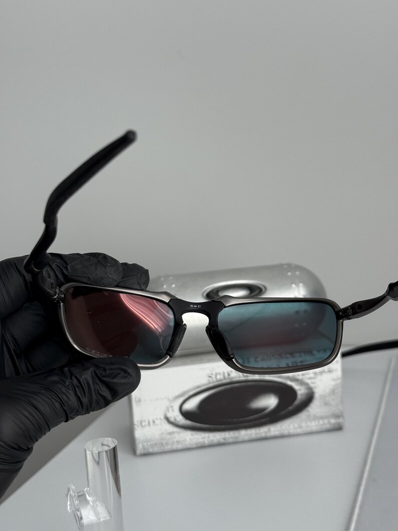 May include: Black rectangular sunglasses with a silver frame and gradient lenses. The lenses transition from red to blue. The sunglasses are held in a black gloved hand. A silver case with the brand name is in the background.