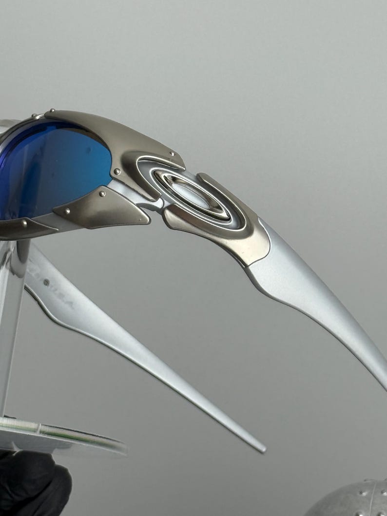 May include: Silver-framed sunglasses with blue lenses. The sunglasses have a sleek, modern design with a metallic finish. The lenses are a vibrant blue, contrasting with the silver frame. The arms of the sunglasses are long and curved.
