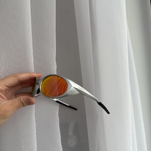 May include: Silver-framed sunglasses with orange lenses. The sunglasses have a sleek, modern design with black earpieces. The lenses are a vibrant orange color, offering a bold contrast to the silver frame.