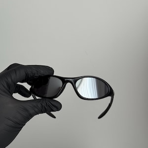 K&ouml;nnte beinhalten: Schwarze Oakley-Sonnenbrille mit gebogenem Rahmen und dunklen Gl&auml;sern. Die Sonnenbrille wird von einer behandschuhten Hand vor einem schlichten Hintergrund gehalten. Das Oakley-Logo ist oben auf dem Rahmen sichtbar.