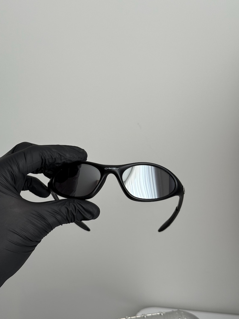 K&ouml;nnte beinhalten: Schwarze Oakley-Sonnenbrille mit gebogenem Rahmen und dunklen Gl&auml;sern. Die Sonnenbrille wird von einer behandschuhten Hand vor einem schlichten Hintergrund gehalten. Das Oakley-Logo ist oben auf dem Rahmen sichtbar.