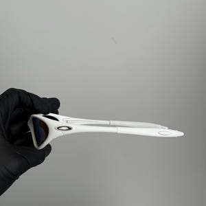 May include: White sports sunglasses with a sleek, modern design. The sunglasses have a white frame and temples, with dark lenses. The glasses are held by a gloved hand against a plain grey background.
