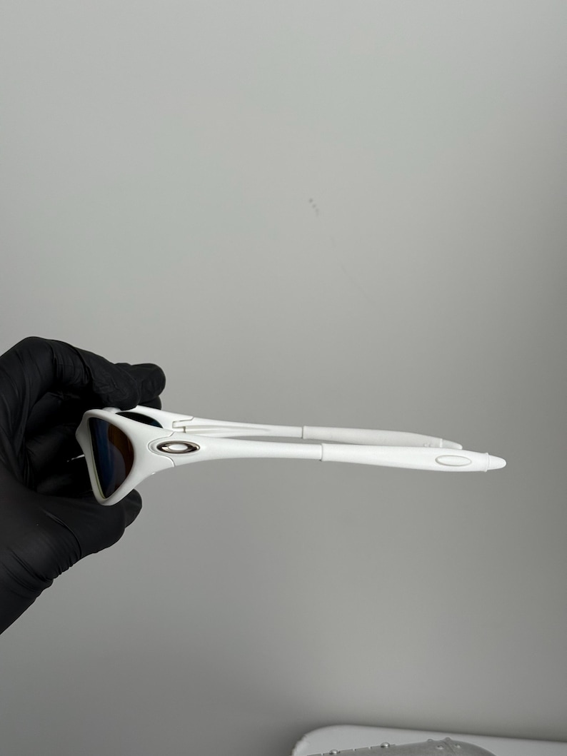 May include: White sports sunglasses with a sleek, modern design. The sunglasses have a white frame and temples, with dark lenses. The glasses are held by a gloved hand against a plain grey background.