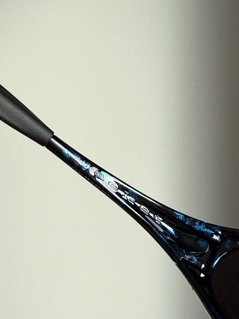 May include: Close-up of a black and blue patterned sunglasses arm. The arm has a glossy finish with blue accents and the word "Police" printed on the side. The sunglasses arm is set against a light background.