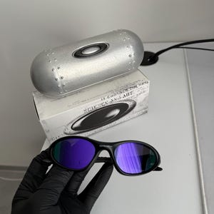 May include: Black sunglasses with purple mirrored lenses. The sunglasses are held by a gloved hand. A silver case and a box with the text "SCIENCE AND ART" are in the background.