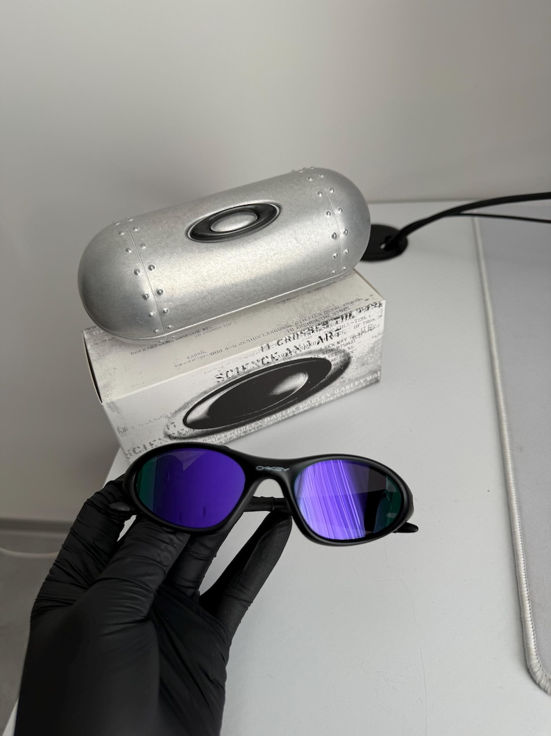 May include: Black sunglasses with purple mirrored lenses. The sunglasses are held by a gloved hand. A silver case and a box with the text "SCIENCE AND ART" are in the background.