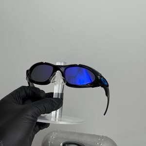 May include: Black sunglasses with blue reflective lenses. The sunglasses are held on a clear display stand. A silver case with a black oval logo is below the sunglasses.