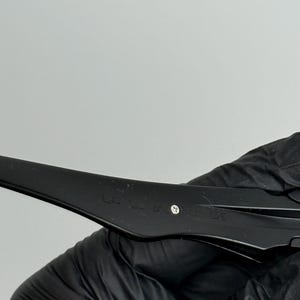 May include: Close-up of a black object with a curved design. The object has a matte finish and appears to be made of plastic or a similar material. A small, silver-colored rivet is visible. The object is held by a gloved hand.