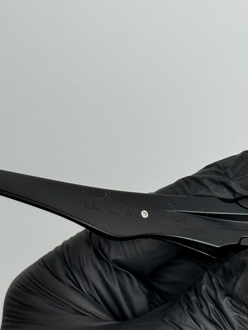 May include: Close-up of a black object with a curved design. The object has a matte finish and appears to be made of plastic or a similar material. A small, silver-colored rivet is visible. The object is held by a gloved hand.