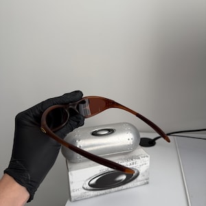 May include: Brown and tan sunglasses with a matching case and box. The sunglasses have a curved frame and dark lenses. The case is silver and the box is white with black text and a logo.