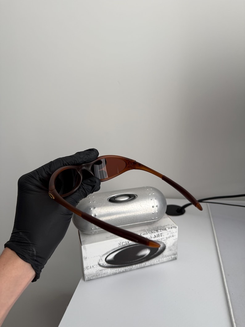 May include: Brown and tan sunglasses with a matching case and box. The sunglasses have a curved frame and dark lenses. The case is silver and the box is white with black text and a logo.