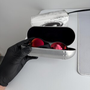 May include: Red-lensed sunglasses in a silver case. The sunglasses have a black frame and are in an open, silver case. The case is held by a gloved hand. The lenses reflect light. The case has a black interior.