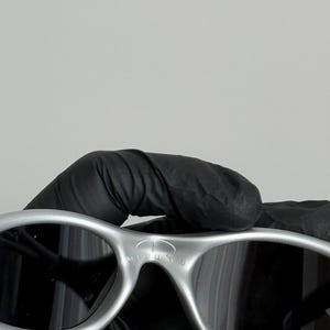 Puede incluir: Gafas de sol plateadas con lentes oscuras. Las gafas tienen un dise&ntilde;o curvo y el texto "1 MINUTE" est&aacute; impreso en la montura. Las gafas de sol est&aacute;n sujetas por una mano enguantada.