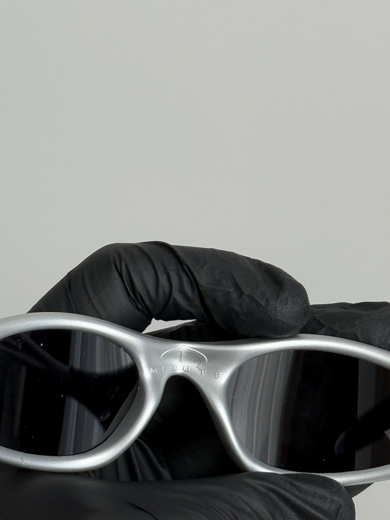 Puede incluir: Gafas de sol plateadas con lentes oscuras. Las gafas tienen un dise&ntilde;o curvo y el texto "1 MINUTE" est&aacute; impreso en la montura. Las gafas de sol est&aacute;n sujetas por una mano enguantada.