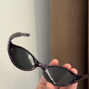 May include: Oval-shaped sunglasses with dark lenses and a purple and black patterned frame. The sunglasses have a curved design and are held in a hand.