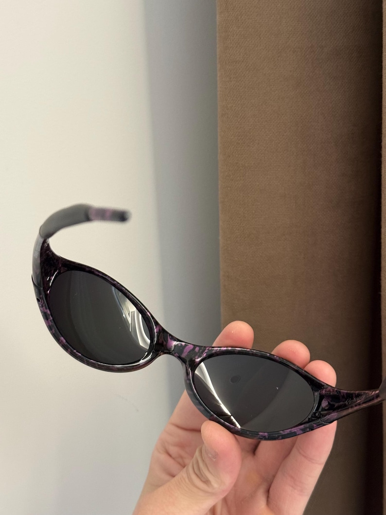 May include: Oval-shaped sunglasses with dark lenses and a purple and black patterned frame. The sunglasses have a curved design and are held in a hand.