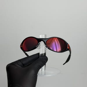 May include: Black sunglasses with red lenses and orange accents on the temples. The sunglasses are held on a clear display stand. The lenses reflect a purple hue.